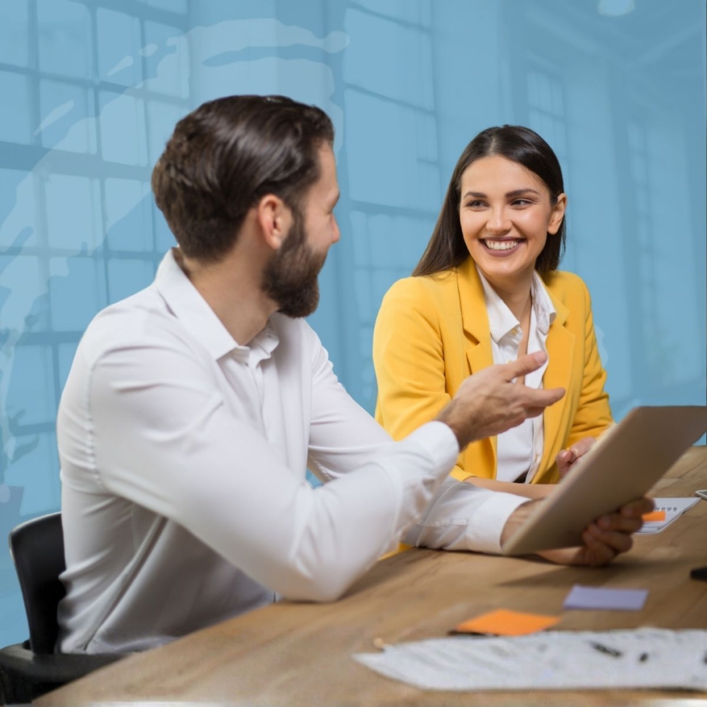 Two colleagues at a wooden table discuss with a tablet, smiling in a bright office setting.