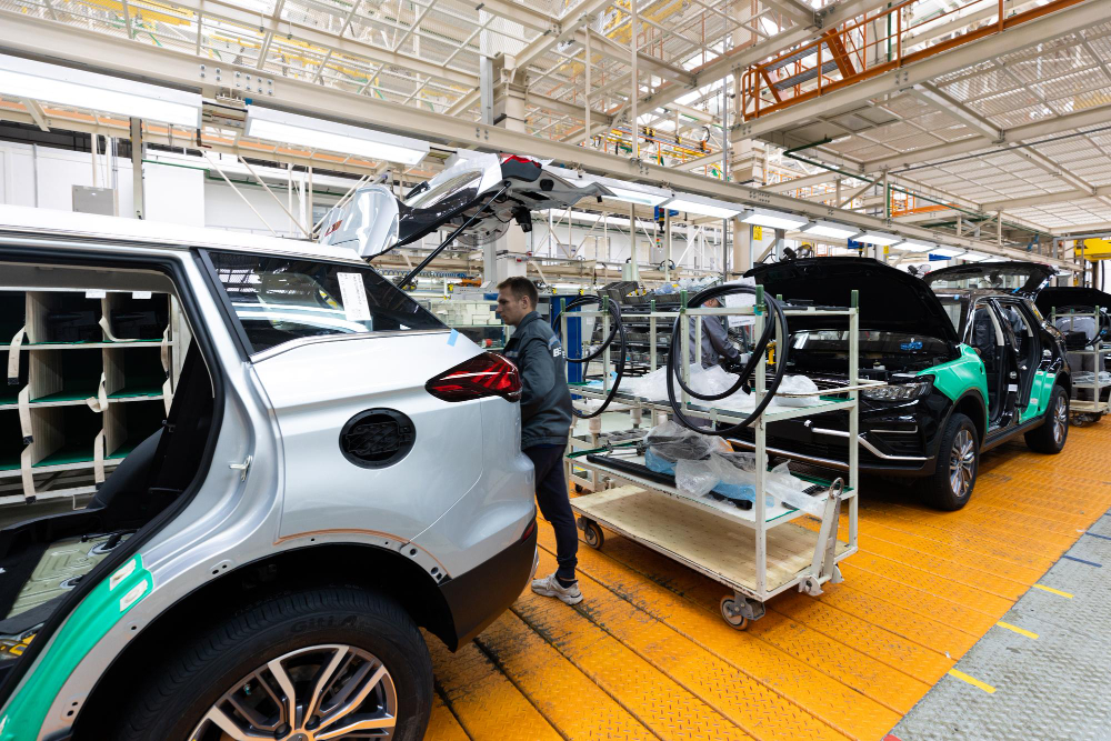 Automotive factory assembly line with a silver SUV having its rear hatch open as a technician works nearby and a cart of parts sits on the orange factory floor.